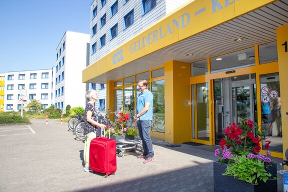 Eine Rehabilitandin reist mit einem roten Koffer an. Sie wird von einem Mitarbeitenden vor dem Klinikeingang begrüßt. 