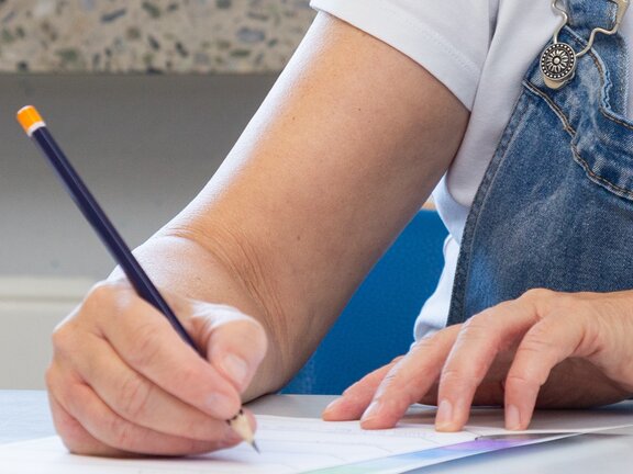 Eine Person schreibt mit einem Bleistift auf ein Blatt an einem Tisch.