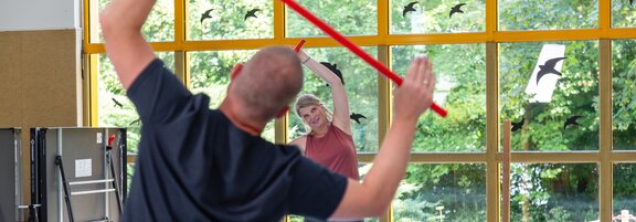 Zwei Rehabilitanden machen Dehnübungen in einer Sporthalle. Im Hintergrund ist ein großes Fenster. Der Blick geht direkt ins Grüne. 