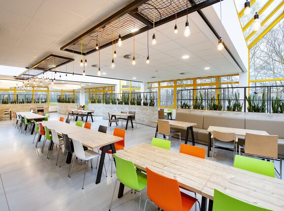 Die Cafeteria ist sehr hell und freundlich gestaltet. Die Stühle haben unterschiedliche Farben. 