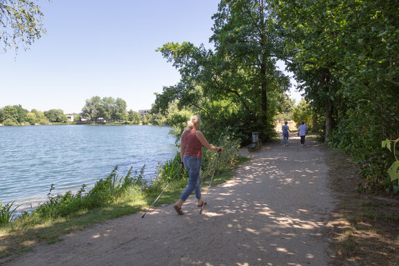 Drei Rehabilitandinnen gehen mit Nordic-Walking-Stöcken um den See. Die Sonne scheint. Das Wasser im See ist blau und im Hintergrund sieht man 4 kleine Enten. 