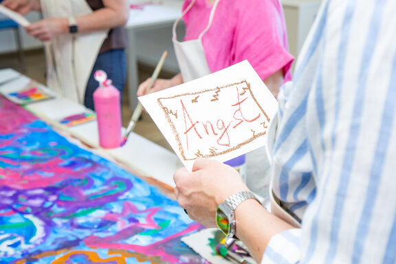 Eine Frau steht in einem Kunstraum und hält ein Schild in der Hand. Auf diesem Schild steht das Wort Angst.