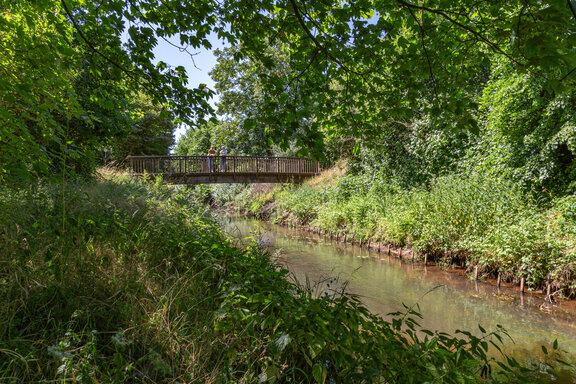 Bild einer Brücke, die über einen Fluss geht. Zwei Rehabilitandinnen stehen bei schönem Wetter an der Brücke und schauen auf den Fluss.