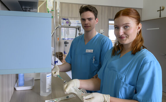 Zwei HPA Auszubildene stehen an einem Tisch und schauen in die Kamera. Beide tragen Handschuhe und haben eine Spritze in der Hand. 
