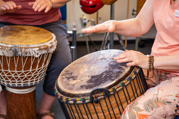 Auf dem Bild sitzen zwei Rehabilitandinnen nebeneinander und trommeln auf einem Djembem (eine afrikanische Trommel, die hauptsächlich mit den Händen gespeilt wird.). 