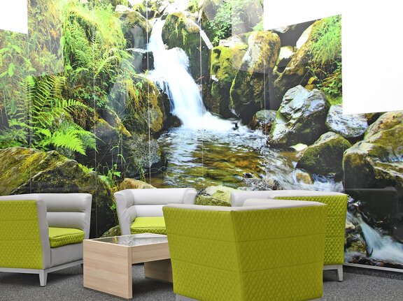 Vier grüne gemütliche Sessel stehen vor einem rieseigen Glas Bild auf dem ein Wasserfall abgebildet ist. 