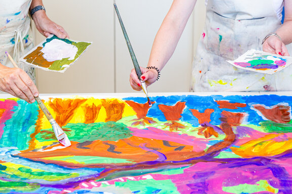 Zwei Rehabilitanden bemalen eine große Leinwand mit bunten Farben. 