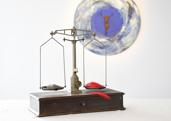 Wir sehen eine antike Waage abgebildet. In der einen Schale liegt ein Stein in der anderen liegt eine Feder und dennoch halten Sie sich die Waage. Im Hintergrund sehen wir ein Kreuz.