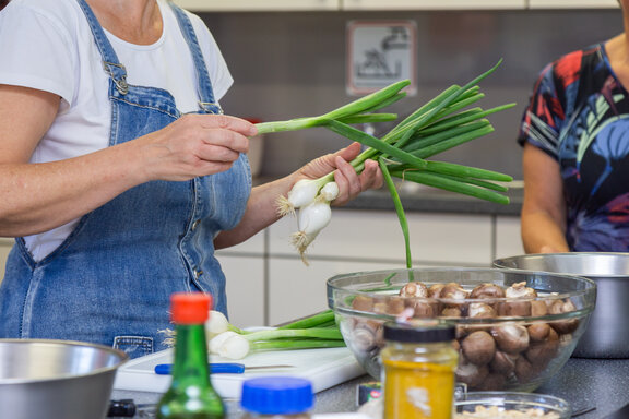 Eine Rehabilitandin schneidet in der Lehrküche Frühlingszwiebeln. Drumherum stehen auf der Kochinsel weitere Zutaten und im Hintergrund ist eine weitere Person im Anschnitt zu sehen.