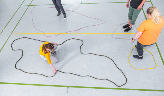 Drei Rehabilitand*innen und eine Therapeutin legen farbige Seile auf dem Fußboden aus. Sie legen die Seile aus, dass sie sich in der geschlossenen Form des Kreises befinden. 