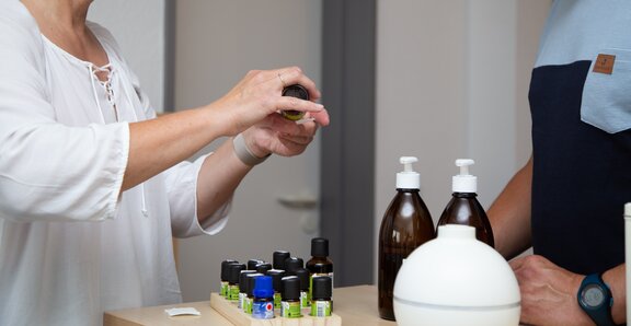 Eine Frau tropft Aromaöl auf einen Tupfer. Ein Rehabilitand ist bei ihr. Vor ihnen stehen verschiedene Aromaöle und ein Vernebler. 