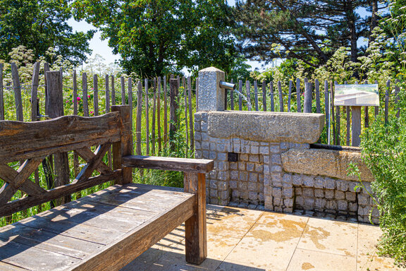 Inmitten von Kräutern und Bäumen steht ein Staketenzaun. Im Inneren befindet sich an der rechten Seite eine massive Holzbank. Geradeaus befindet sich das Unterarmwasserbecken. Es ist aus groben Steinen gemauert. Im oberen Bereich läuft Wasser ins Becken.