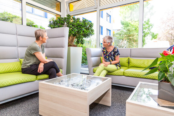 Zwei Rehabilitandinnen unterhalten sich. Sie sitzen in einer grau-grünen Sitzecke im Foyer. 