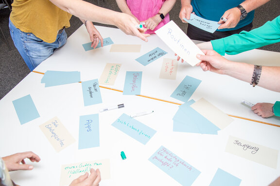 Eine Therapiegruppe hat sich um einen Tisch verteilt und schreibt Stichworte auf farbige Karten. 