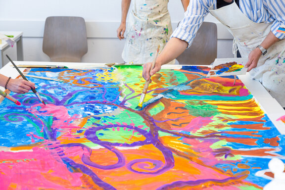 Mehrere Rehabilitanden bemalen eine große Leinwand mit bunten Farben. 