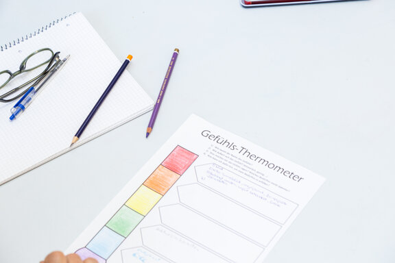 Es liegen ein paar Schreibutensilien auf einem Tisch. Im Vordergrund liegt ein "Gefühlsbarometer". 